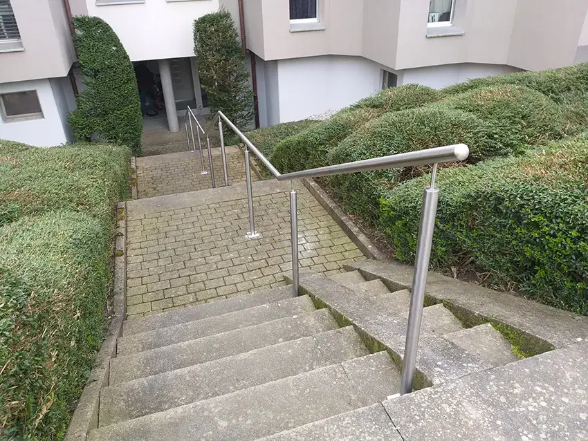 Main courante en inox sur un escalier extérieur pavé, entre des haies